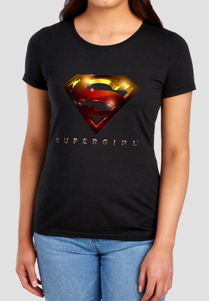 Woman wearing black t-shirt with shiny gold and red Supergirl logo and text, paired with light blue jeans against a gray background.