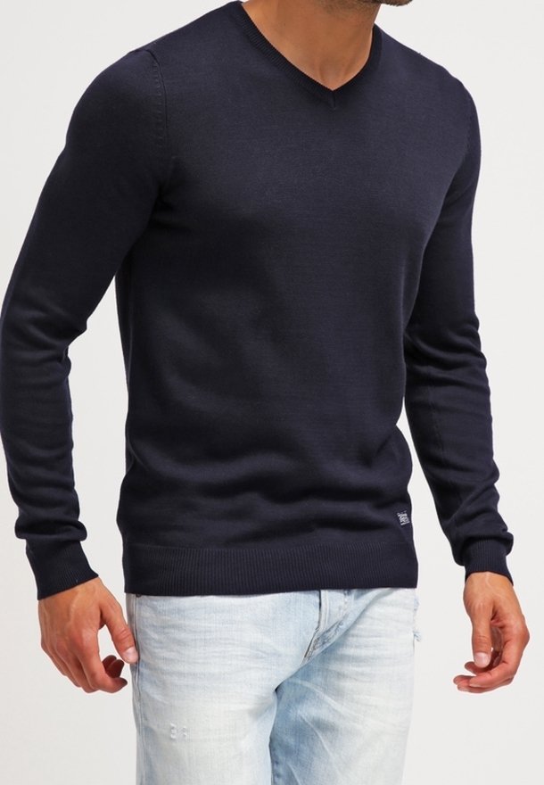 Pull tricot bleu marine avec col en V, manches longues et poignets et ourlet côtelés. Porté avec un jean bleu clair, mettant en valeur une coupe ajustée.