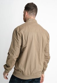 Man wearing a light brown casual jacket and dark jeans, standing with his back to the camera against a plain white background.