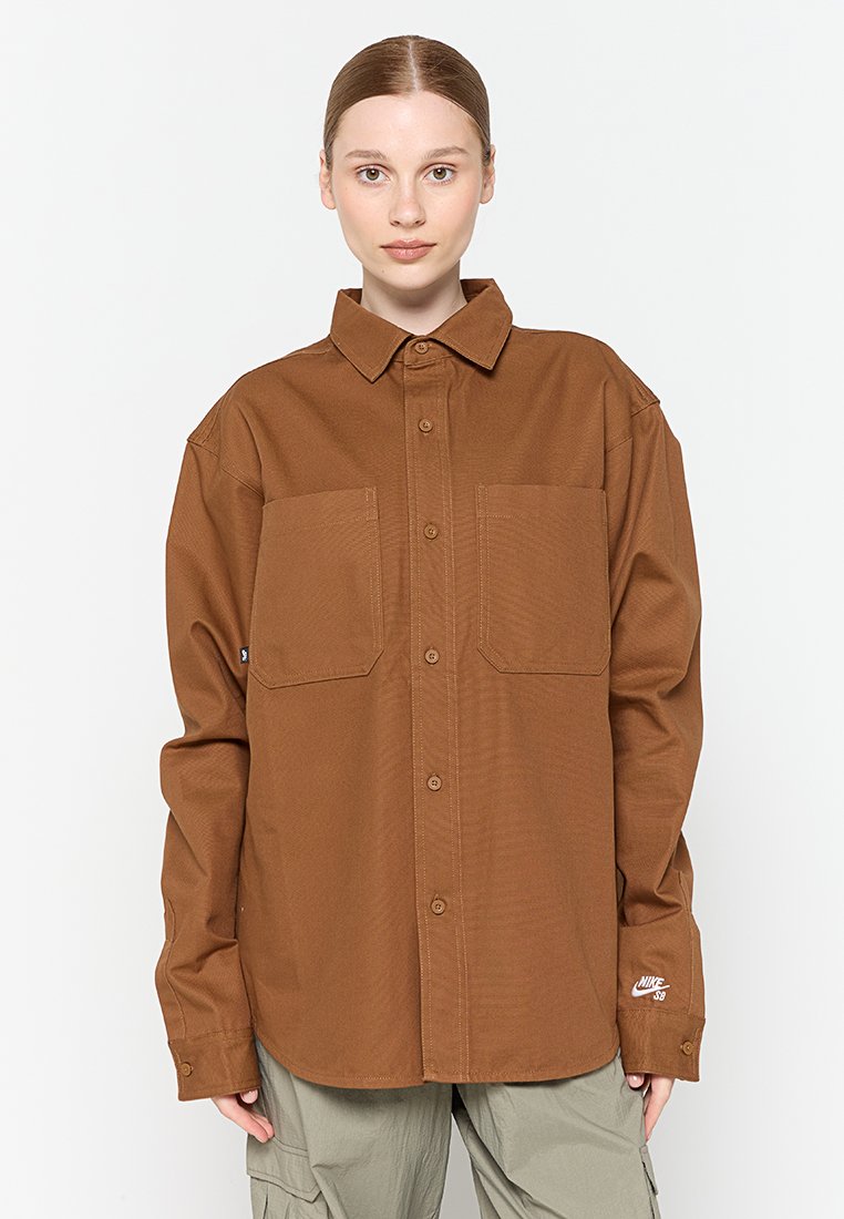 Chemise marron à boutons avec deux poches avant, manches longues et un petit logo sur l'ourlet. Le tissu a une texture lisse.