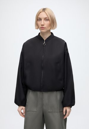 Jeune personne aux cheveux blonds courts portant une veste noire zippée et un pantalon gris, debout sur un fond blanc uni.