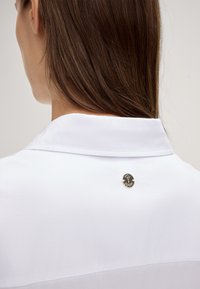Witte button-up overhemd met een puntkraag, voorzien van een goudkleurige ronde logo-pin op de achterkant. Glad textuur, minimalistisch ontwerp.