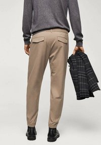 Pantalon beige avec poignets élastiques, deux poches arrière, assorti à une veste noire à carreaux. Le tissu semble lisse et structuré.