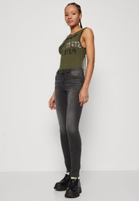 Olivenfarget ribbestrikket bodysuit med grafisk trykk, kombinert med slim-fit mørkegrå jeans. Modellen har på seg klumpete svarte sko.