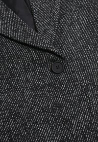 Svart- och vitprickigt tyg med en texturerad yta, med en svart knapp och skårad kavajslagdetalj på en skräddarsydd kavaj.