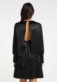 Sort satin kjole med lav ryg og snørelukning, lange ærmer og knaplukninger. Glat overflade med et flydende design.