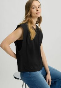 Černý bezrukávový top s širokými průramky, vyrobený z měkkého materiálu, se zdobným detailem na lemu. V kombinaci s modrými džíny.