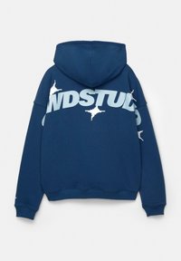 Tummansininen huppari, jossa on suuri valkoinen "STUDS"-teksti, tekstuuriset yksityiskohdat ja pudotetut olkaimet. Pehmeä, puuvillasekoitekangas.