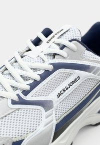 Adidași albi și navy, cu partea superioară din plasă, accente sintetice, logo proeminent și talpă texturată. Prezintă șireturi albe și guler căptușit.
