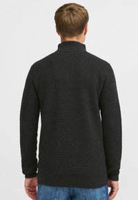 Strikket svart genser med høyt hals, teksturert overflate og ribbestrikkede ermer og nederkant, vist fra baksiden.