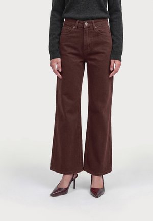 Femme portant un pantalon large brun foncé, un pull gris foncé et des talons hauts pointus foncés, debout devant un fond uni.