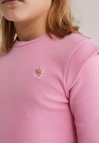 Tricou roz cu mâneci lungi, cu dungi, având o broderie mică cu floare albă și roz pe partea stângă a pieptului. Textură netedă și design ajustat.