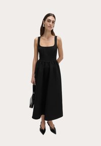Schwarzes Spaghetti-Trägerkleid mit quadratischem Ausschnitt, tailliertem Oberteil und ausgestelltem Rock. Das Model hält eine schwarze Handtasche und trägt spitze schwarze High Heels.