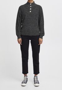 Person in einem dunkelgrauen Strickpullover mit vier weißen Knöpfen, schwarzen cropped Hosen und schwarzen High-Top-Sneakers mit weißen Schnürsenkeln.