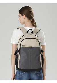Zaino in tessuto beige e grigio, con chiusura a doppia zip, spallacci regolabili e una tasca frontale con dettagli di cucitura marrone.