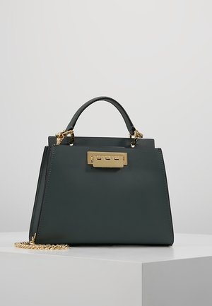 Donkergroene trapeziumvormige handtas met handvat bovenop, gouden sluiting en afneembare gouden kettingband op een wit oppervlak tegen een effen grijze achtergrond.