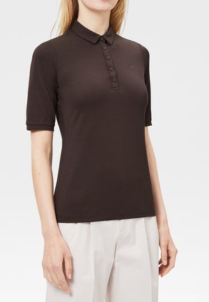 Kobieta ubrana w dopasowaną brązową koszulkę polo z krótkim rękawem i plisą zapinaną na guziki, w połączeniu z luźnymi białymi spodniami, na jasnym jednolitym tle.