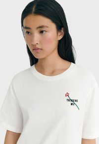 Junge Frau mit langen dunklen Haaren, die ein weißes T-Shirt mit einer kleinen roten Blume und dem Text "THINKING MU" auf der Brust trägt.
