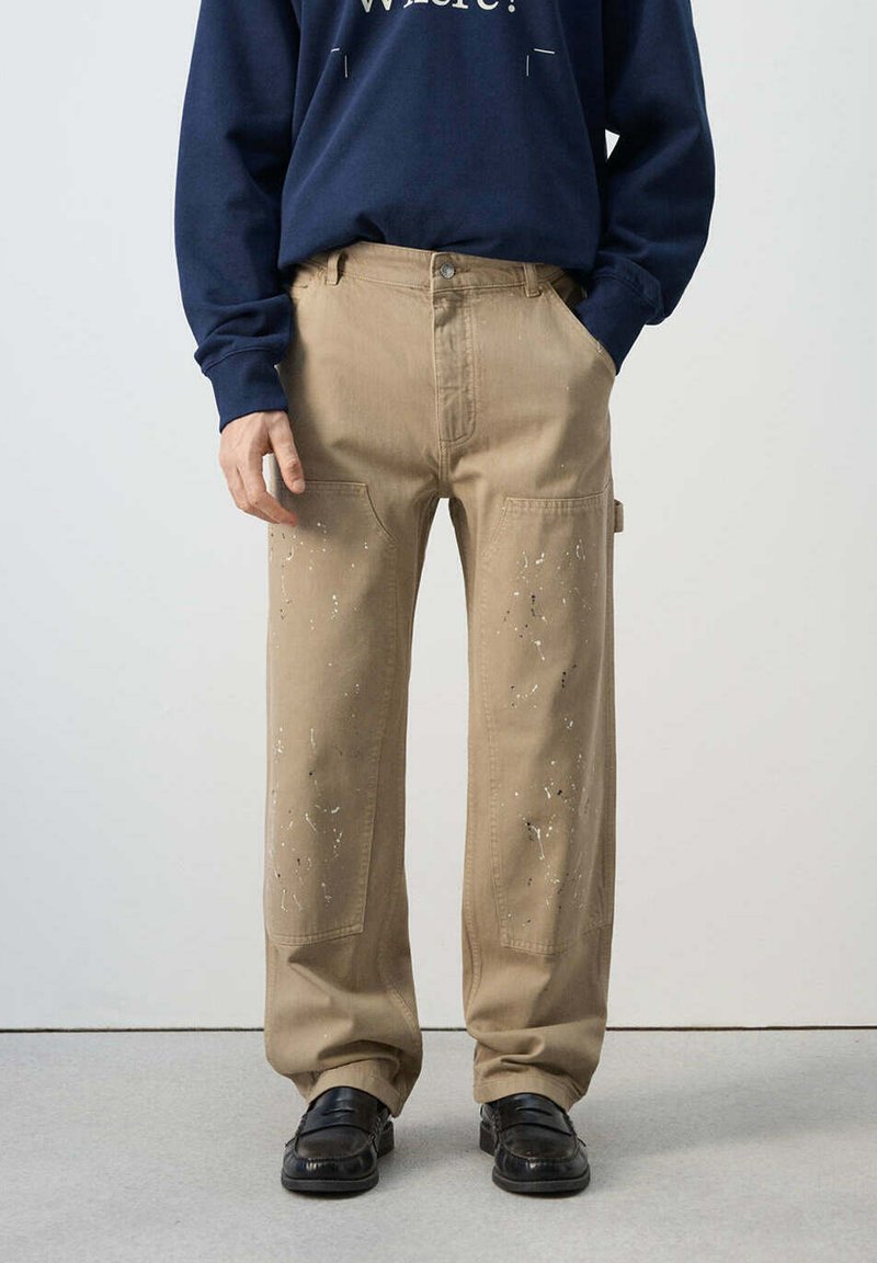 Hombre que lleva pantalones de trabajo beige salpicados de pintura con grandes bolsillos delanteros, zapatos negros y una sudadera azul marino con texto blanco.