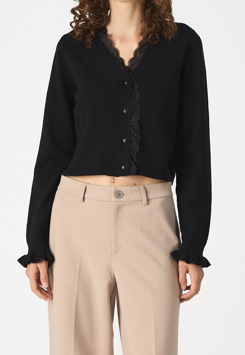 Femme portant un cardigan court noir avec bordure en dentelle et boutons assortis, associé à un pantalon beige taille haute avec fermeture à bouton.