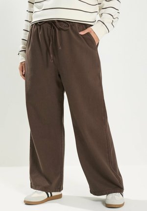 Person trägt braune, weit geschnittene Hose mit Kordelzug, weiße Turnschuhe mit schwarzen Streifen und ein langärmeliges weißes Oberteil mit braunen Streifen.