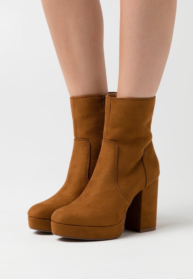 Bottes compensées en suède marron mi-mollet avec talons carrés portées sur des jambes nues sur un fond neutre.