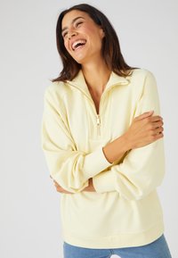 Sudadera de felpa amarilla claro con cuello de media cremallera y acentos acanalados. Textura suave y corte relajado, combinada con unos jeans azules.