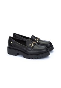 Mocasines de cuero negro con suela de goma gruesa, que cuentan con una hebilla decorativa dorada y detalles de costura. Textura suave con diseño de calzado sin cordones.