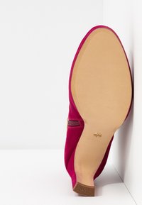 Chaussure à talon haut en daim rouge unique vue par le dessous, mettant en valeur la semelle lisse et le talon carré avec le logo de la marque embossé.
