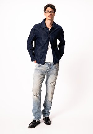 Ung mand iført sorte briller, marineblå stribet jakke, hvid T-shirt, slidte lyseblå jeans og sorte loafers, stående foran en hvid baggrund.