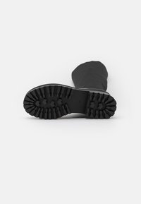 Bota de goma negra con una suela texturizada que presenta profundas ranuras para tracción. La parte superior tiene un diseño liso y está ligeramente caída.