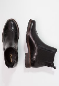 Botas negras de cuero hasta el tobillo con detalles de brogue, paneles laterales elásticos y una suela de goma marrón. Textura suave, diseño elegante y acentos en contraste.