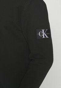 Juodos ilgomis rankovėmis drabužio rankovė su stačiakampiu pleistru, kuriame baltai parašyta „cK“ ir „Calvin Klein Jeans“.