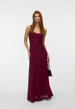 Femme en robe maxi bordeaux en dentelle sans manches tenant un petit sac à main noir sur fond clair uni.