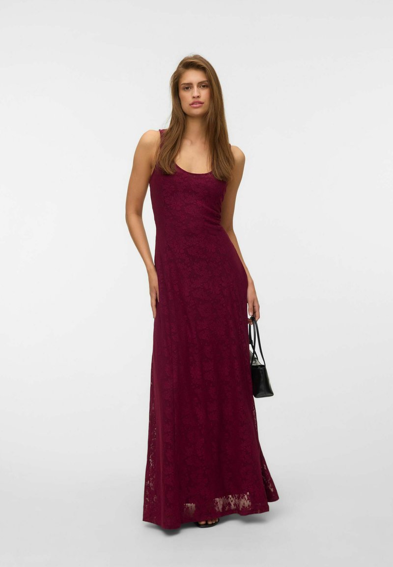 Femme en robe maxi bordeaux en dentelle sans manches tenant un petit sac à main noir sur fond clair uni.