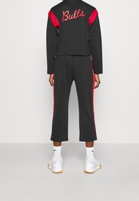 Sort træningsdragt med røde detaljer, der har "Bulls" broderet på ryggen. Toppen er cropped, og bukserne er bredde ben med sidelange striber.