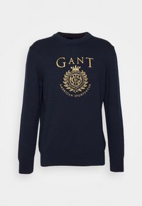 Granatowy sweter dziany z dekoltem w łódkę i długimi rękawami, z złotym haftowanym napisem i logo na środku. Mankiety i dół wykończone ściągaczem.