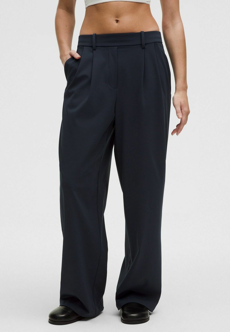 Frau, die hochgeschnittene, lässig geschnittene schwarze Hose mit Taschen trägt, kombiniert mit schwarzen Schuhen, steht vor einem schlichten hellgrauen Hintergrund.