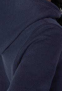 Tejido de sudadera de color azul marino con una textura suave, que presenta un primer plano de la costura del hombro y la capucha. Diseño simple y funcional.