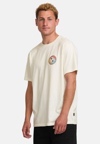 Cream-colored cotton t-shirt with short sleeves and a round neck, featuring a circular logo on the chest in orange, blue, and black.