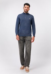 Granatowy sweter zapinany na zamek, tekstura dzianiny, ściągacze przy rękawach i u dołu, złote logo na piersi; w komplecie z szarymi, wyblakłymi dżinsami i beżowymi trampkami.