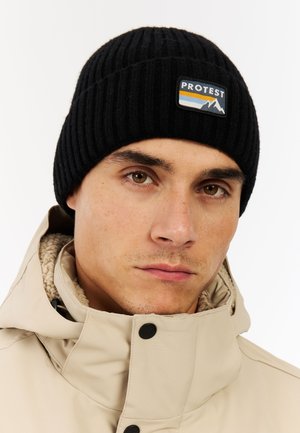 Black ribbed knit beanie with "PROTEST" label patch featuring mountain design. Worn with a beige jacket with a textured collar.