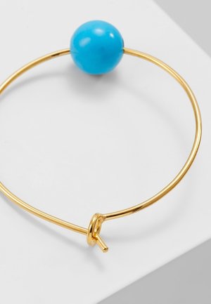 Bracelet jonc en or avec une grande perle turquoise ronde et une fermeture par nœud simple posé sur une surface blanche.