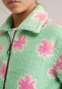 Primo piano di labbra e mento che indossano una giacca in pile verde menta con motivi floreali rosa e una cerniera bronzo, su uno sfondo semplice.