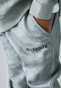 Pilkos sportinės kelnės, pagamintos iš minkštos medžiagos, su šonine kišenę ir juodu "ALLSAINTS" logotipu šalia klešnių, su raišteliais juosmenyje ir ant rankogalio.