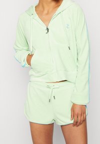 Žena oblečená v světle zelené sametové mikině na zip s předními kapsami a odpovídajícími šortkami s elastickým pasem a modrými detaily na rukávech a šortkách.