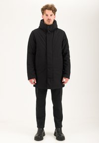 Cappotto imbottito nero con colletto alto, zip frontale e chiusura a scatto, due tasche laterali e una superficie liscia, indossato sopra pantaloni neri e stivali.