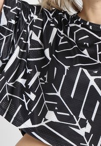 Svart kortärmad topp med ett vitt geometriskt mönster. Materialet har en perforerad struktur som ger andningsförmåga och en lös passform.