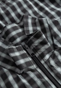 Tissu à carreaux noir et gris avec une fermeture éclair noire partiellement ouverte sur un vêtement ou un textile.