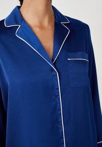 Haut de pyjama en satin bleu avec passepoil blanc, col, fermeture à boutons et poche poitrine gauche. Texture lisse avec une finition brillante.